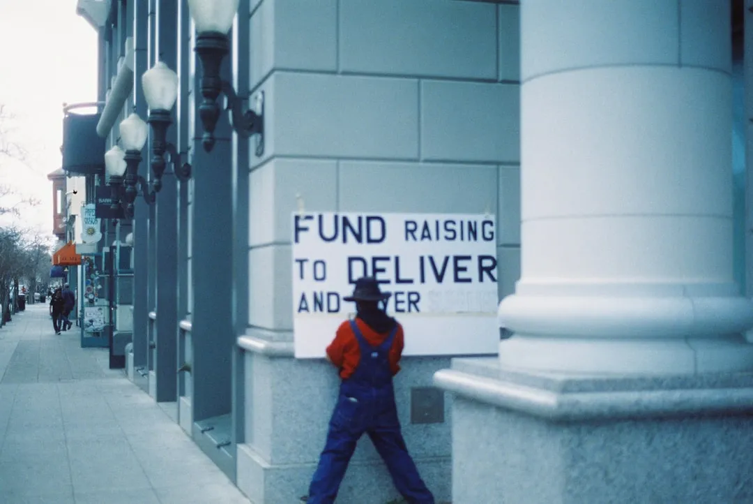 Man in blue jacket and blue denim jeans standing beside a white concrete wall
